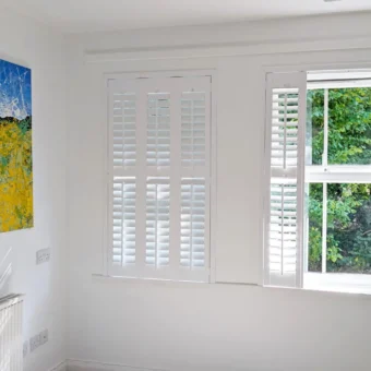 White plantation shutters in a bright Bristol flat, designed by Rob Reeves of Just Shutters. The shutters soften the natural light and frame a leafy woodland view beside a colourful painting.
